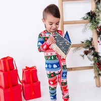 A toddler standing and reading a board book, wearing a zip up pajama in red, green, blue, and white fair isle stripes with snowflakes, pine trees, and white dragon heads.