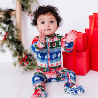 A baby sitting with raised hands and feet forward, wearing a zip up pajama with foot covers in red, green, blue, and white fair isle stripes with snowflakes, pine trees, and white dragon heads.