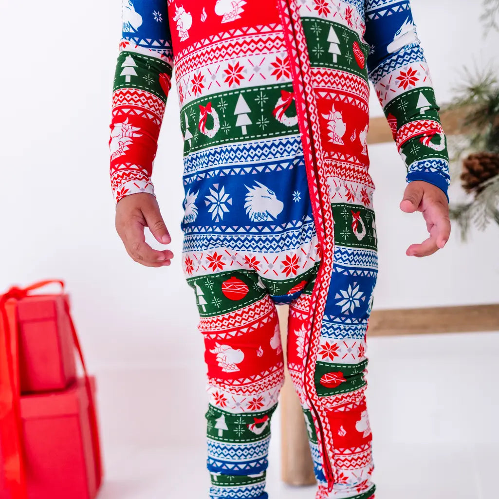 Close up of a standing toddler's torso and legs showing the long front zipper of a pajama in red, green, blue, and white fair isle stripes with snowflakes, pine trees, and white dragon heads.