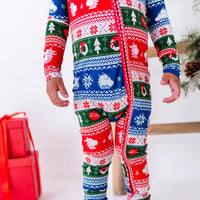 Close up of a standing toddler's torso and legs showing the long front zipper of a pajama in red, green, blue, and white fair isle stripes with snowflakes, pine trees, and white dragon heads.