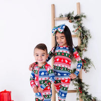 A toddler grinning while an older sibling smiles down, in a matching pajama set and zip up baby pajama in red, green, blue, and white fair isle stripes with snowflakes.