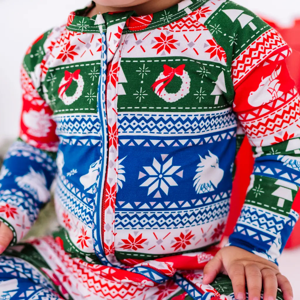 Close up of the front zipper and chest of a zip up baby pajama in red, green, blue, and white fair isle stripes with snowflakes, pine trees, and white dragon heads.