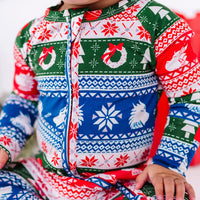 Close up of the front zipper and chest of a zip up baby pajama in red, green, blue, and white fair isle stripes with snowflakes, pine trees, and white dragon heads.