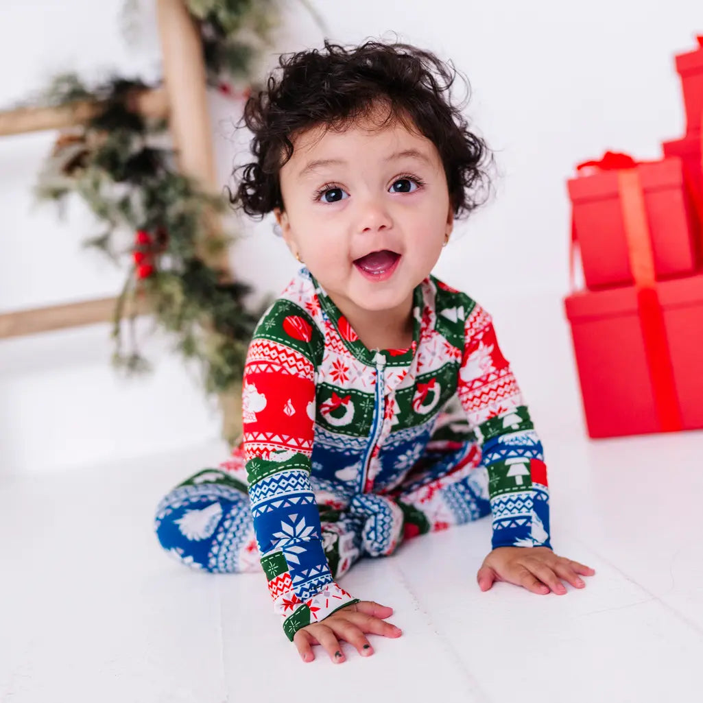 A baby with an open mouth smile leaning forward on both hands, wearing a zip up pajama in red, green, blue, and white fair isle stripes with snowflakes, pine trees, and white dragon heads.