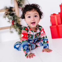 A baby with an open mouth smile leaning forward on both hands, wearing a zip up pajama in red, green, blue, and white fair isle stripes with snowflakes, pine trees, and white dragon heads.