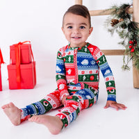 A smiling toddler sitting with legs stretched out and bare feet, wearing a zip up pajama in red, green, blue, and white fair isle stripes with snowflakes, pine trees, and white dragon heads.