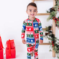 A smiling toddler standing, wearing a zip up pajama with a long front zipper in red, green, blue, and white fair isle stripes with snowflakes, pine trees, and white dragon heads.