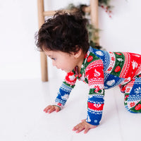 Side view of a baby crawling on a white floor and looking down, wearing a zip up pajama in red, green, blue, and white fair isle stripes with snowflakes, pine trees, and white dragon heads.