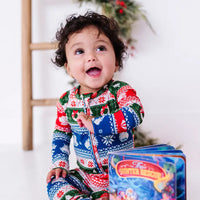 A baby laughing and looking up while sitting next to a board book, wearing a zip up pajama in red, green, blue, and white fair isle stripes with snowflakes, pine trees, and white dragon heads.