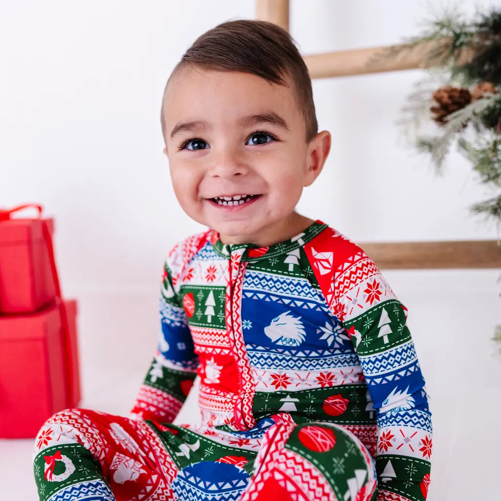 Close portrait of a toddler with a big smile, wearing a zip up pajama in red, green, blue, and white fair isle stripes with snowflakes, pine trees, and white dragon heads.