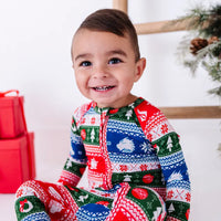 Close portrait of a toddler with a big smile, wearing a zip up pajama in red, green, blue, and white fair isle stripes with snowflakes, pine trees, and white dragon heads.