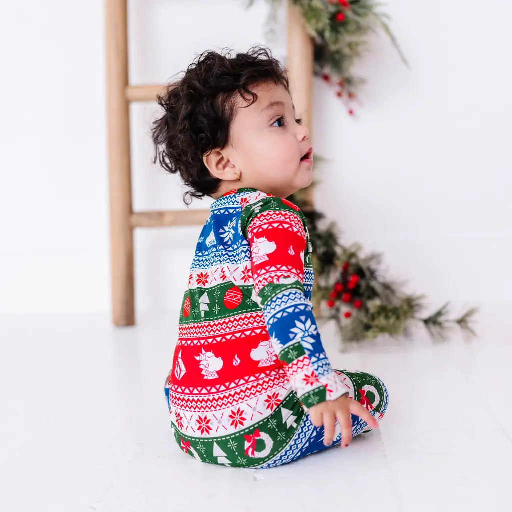 Side view of a baby sitting and looking up, wearing a zip up pajama in red, green, blue, and white fair isle stripes with snowflakes, pine trees, and white dragon heads.