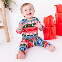 A toddler laughing with raised hands while sitting barefoot, wearing a long sleeve pajama set in red, green, blue, and white fair isle stripes with snowflakes, pine trees, and white dragon heads.