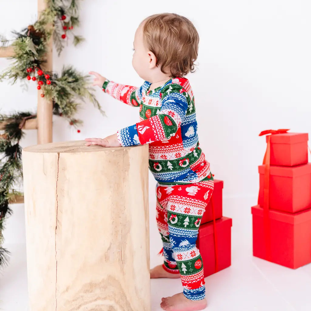 Back view of a toddler reaching toward a garland, wearing a long sleeve pajama set in red, green, blue, and white fair isle stripes with snowflakes, pine trees, and white dragon heads.