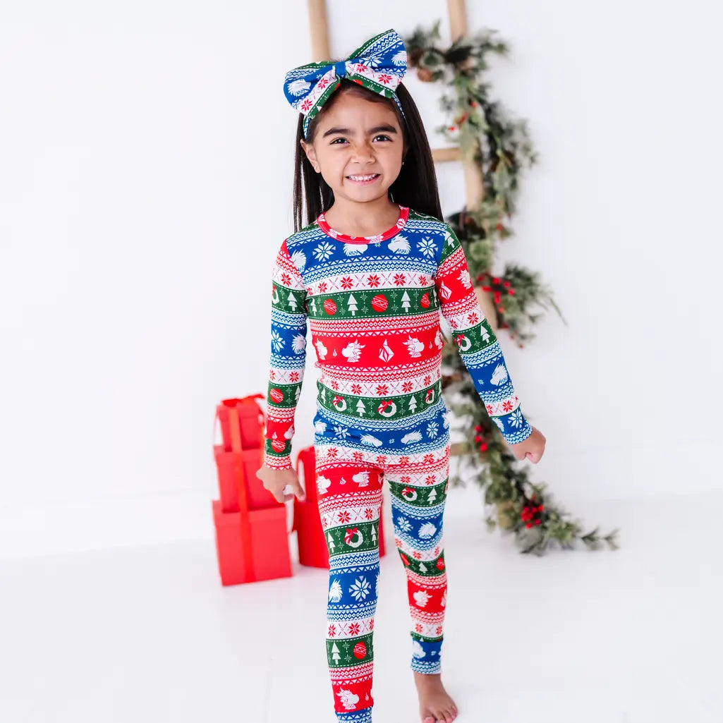 A smiling child in a bow headband walking toward the camera, wearing a long sleeve pajama set in red, green, blue, and white fair isle stripes with snowflakes, pine trees, and white dragon heads.