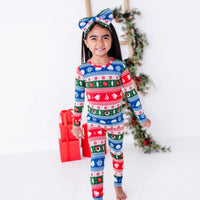 A smiling child in a bow headband walking toward the camera, wearing a long sleeve pajama set in red, green, blue, and white fair isle stripes with snowflakes, pine trees, and white dragon heads.