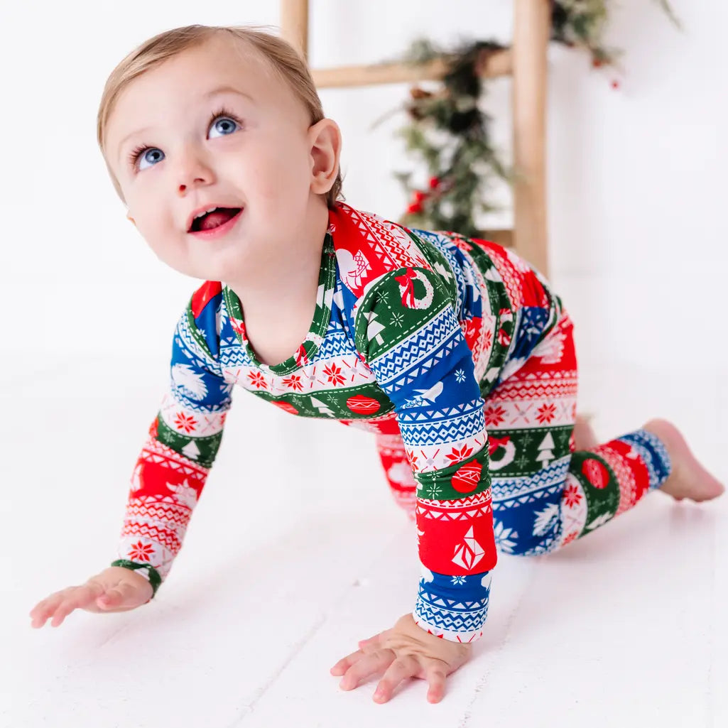 A smiling toddler crawling on a white floor and looking up, wearing a long sleeve pajama set in red, green, blue, and white fair isle stripes with snowflakes, pine trees, and white dragon heads.