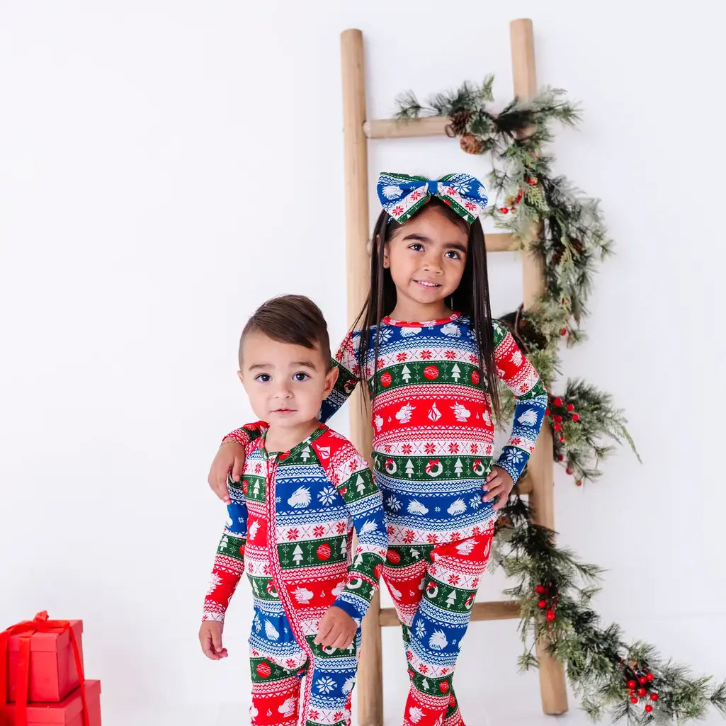Two siblings standing by a ladder and smiling at the camera in a matching pajama set and zip up baby pajama in red, green, blue, and white fair isle stripes with snowflakes.