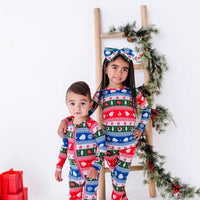 Two siblings standing by a ladder and smiling at the camera in a matching pajama set and zip up baby pajama in red, green, blue, and white fair isle stripes with snowflakes.