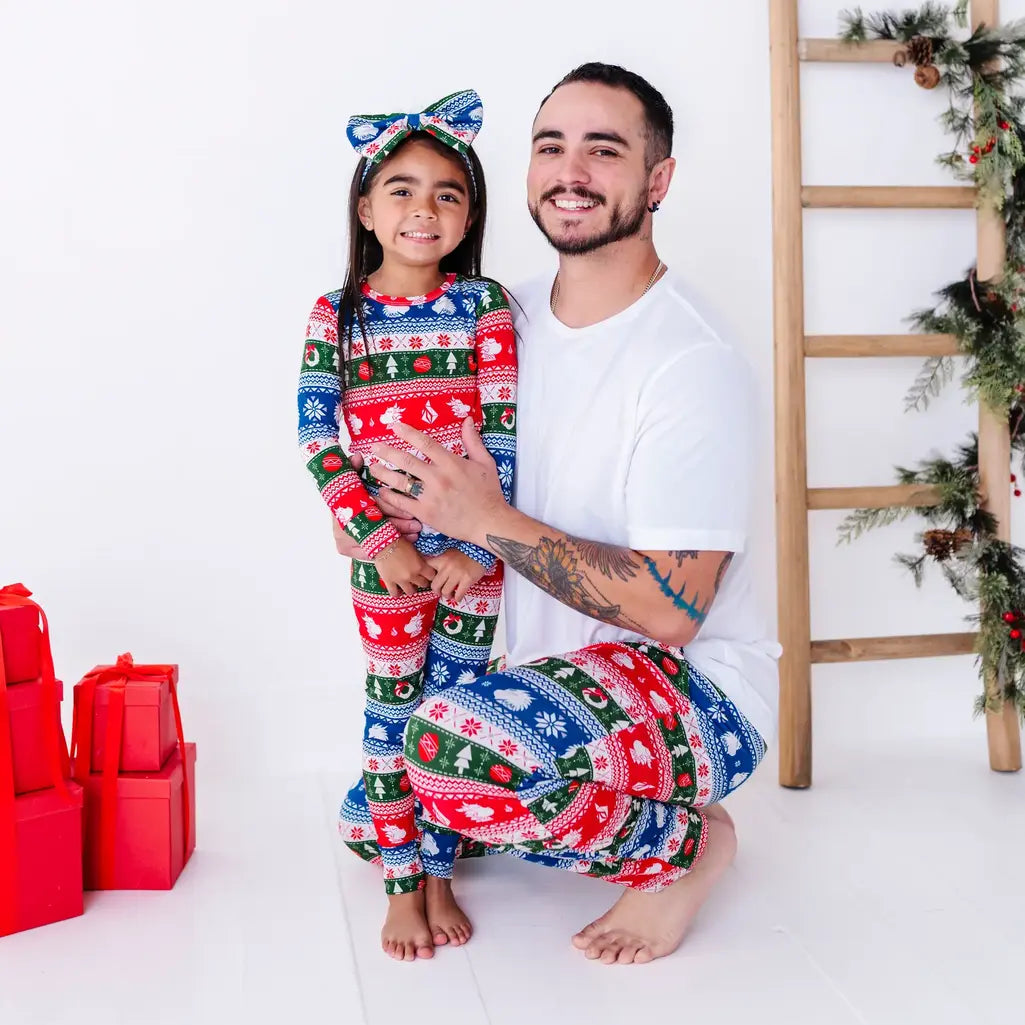 An adult kneeling beside a child, both grinning at the camera in matching pajama pants and a pajama set in red, green, blue, and white fair isle stripes with snowflakes.