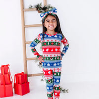 A child grinning with hands on hips, in a bow headband and a long sleeve pajama set in red, green, blue, and white fair isle stripes with snowflakes, pine trees, and white dragon heads.