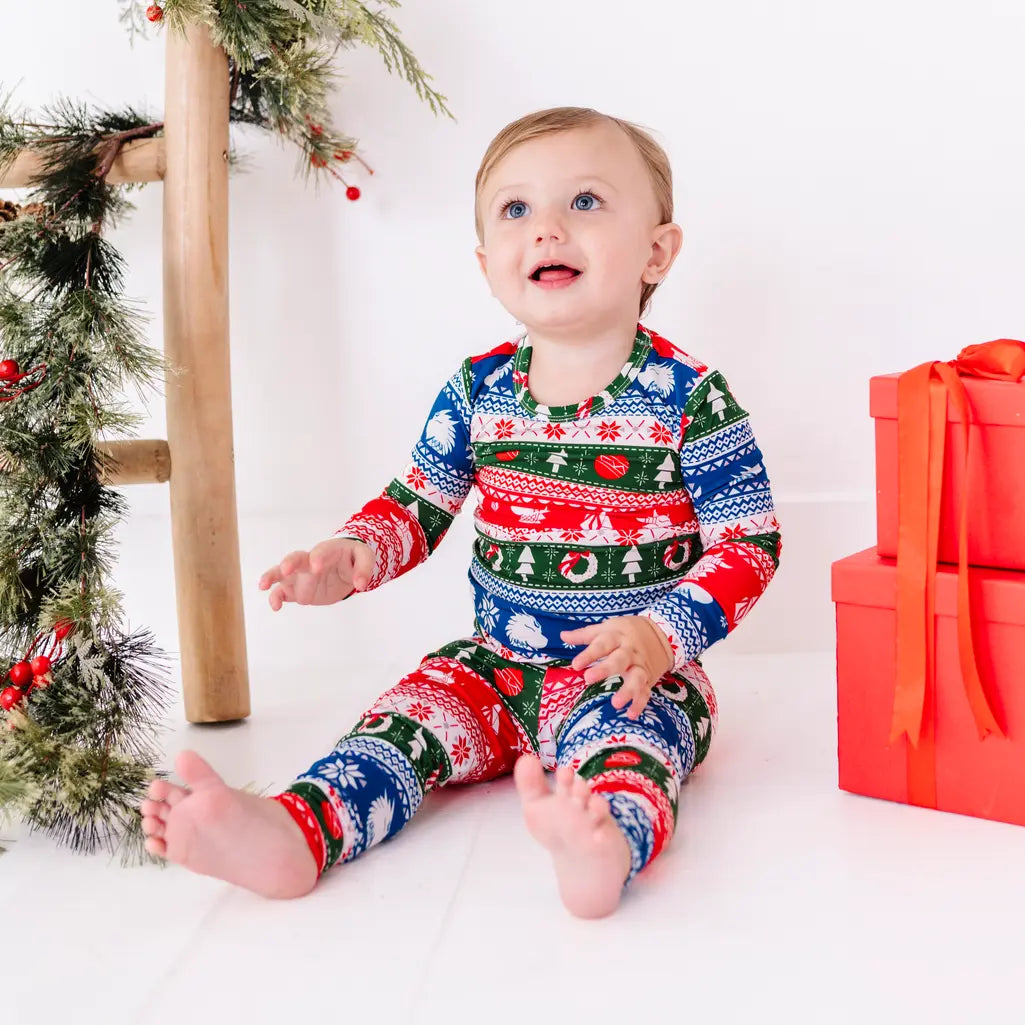 A toddler smiling and looking up while sitting barefoot, wearing a long sleeve pajama set in red, green, blue, and white fair isle stripes with snowflakes, pine trees, and white dragon heads.