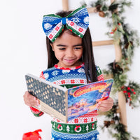 A child in a bow headband smiling down at an open board book, wearing a long sleeve pajama top in red, green, blue, and white fair isle stripes with snowflakes, pine trees, and white dragon heads.