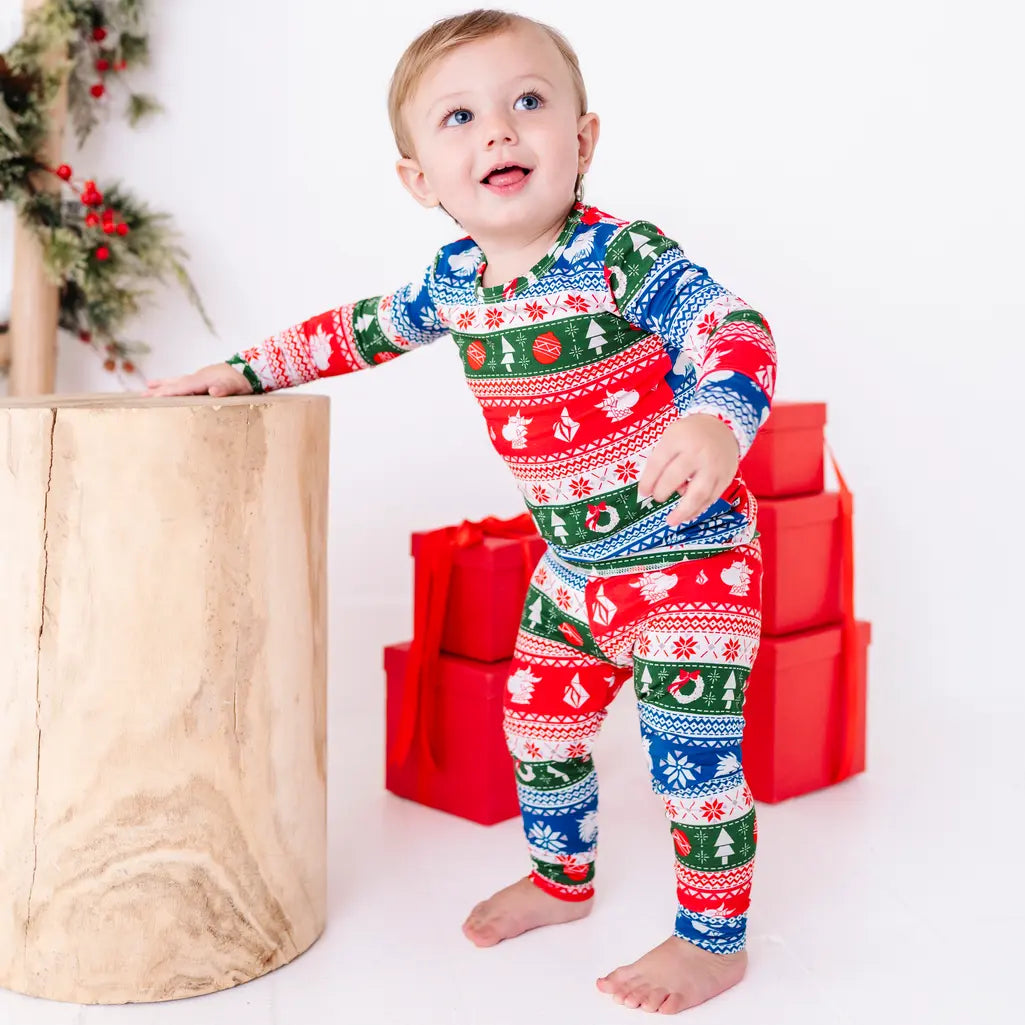 A smiling toddler stepping forward with a hand on a stump, wearing a long sleeve pajama set in red, green, blue, and white fair isle stripes with snowflakes, pine trees, and white dragon heads.