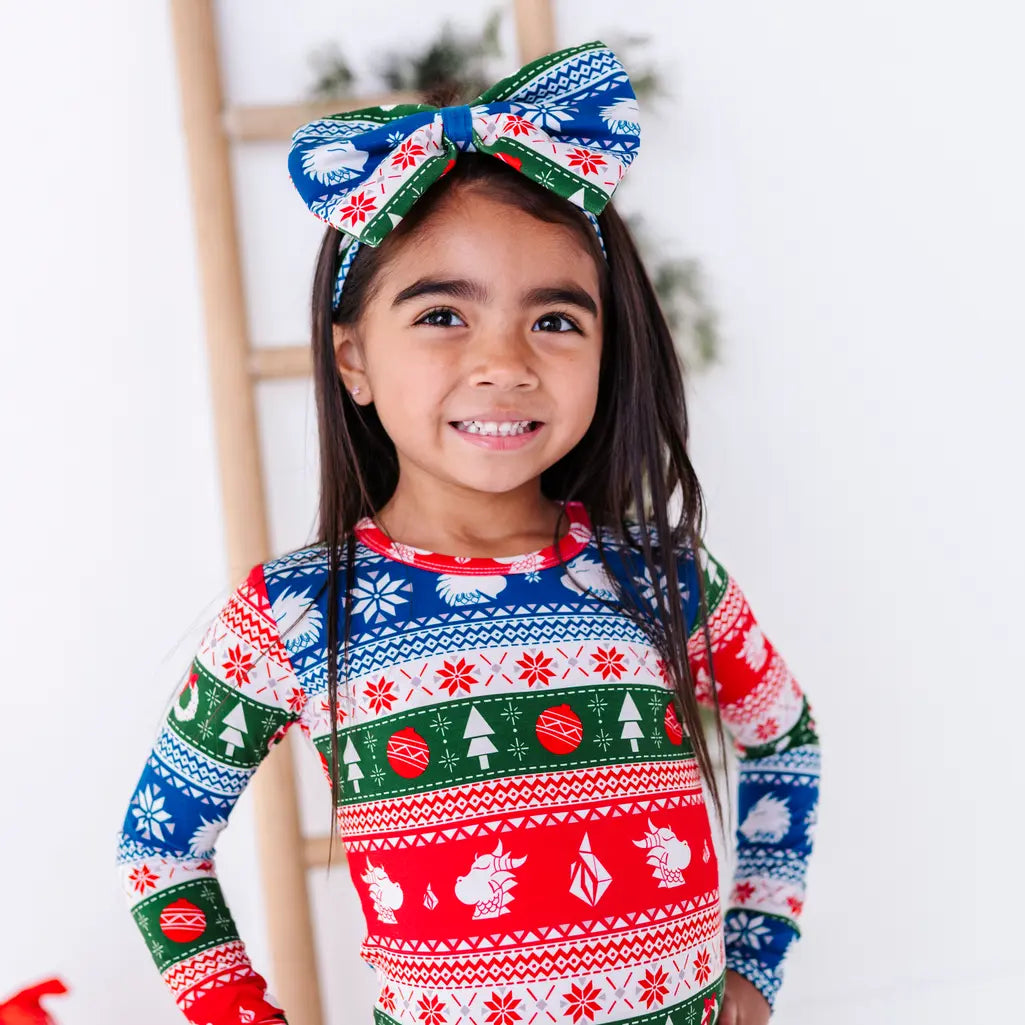 Portrait of a child with a big smile and a bow headband, wearing a long sleeve pajama top in red, green, blue, and white fair isle stripes with snowflakes, pine trees, and white dragon heads.