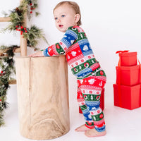 Side view of a toddler leaning on a wooden stump, wearing a long sleeve pajama set in red, green, blue, and white fair isle stripes with snowflakes, pine trees, and white dragon heads.