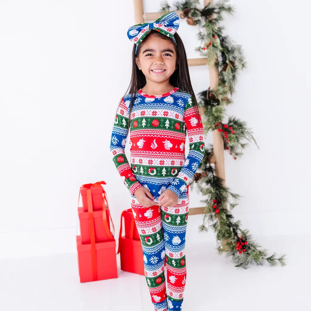 A smiling child in a bow headband standing with hands together, wearing a long sleeve pajama set in red, green, blue, and white fair isle stripes with snowflakes, pine trees, and white dragon heads.