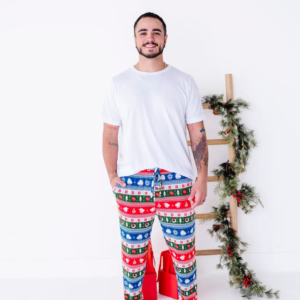 A smiling man in a white tee standing with a hand in the pocket of jogger pajama pants in red, green, blue, and white fair isle stripes with snowflakes, pine trees, and white dragon heads.