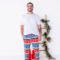 A smiling man in a white tee standing with a hand in the pocket of jogger pajama pants in red, green, blue, and white fair isle stripes with snowflakes, pine trees, and white dragon heads.