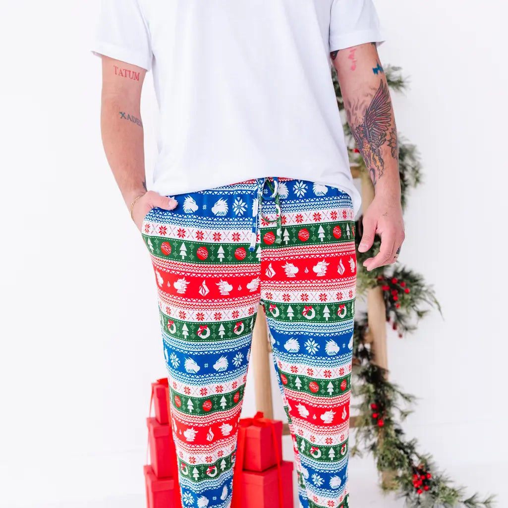 A man with one hand in the pocket, shown from the chest down, wearing jogger pajama pants in red, green, blue, and white fair isle stripes with snowflakes, pine trees, and white dragon heads.