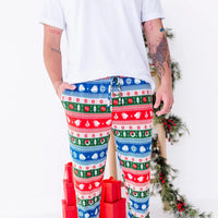 A man with one hand in the pocket, shown from the chest down, wearing jogger pajama pants in red, green, blue, and white fair isle stripes with snowflakes, pine trees, and white dragon heads.