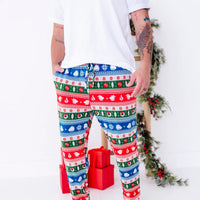 A man with both hands at the pockets, shown from the chest down, wearing jogger pajama pants in red, green, blue, and white fair isle stripes with snowflakes, pine trees, and white dragon heads.