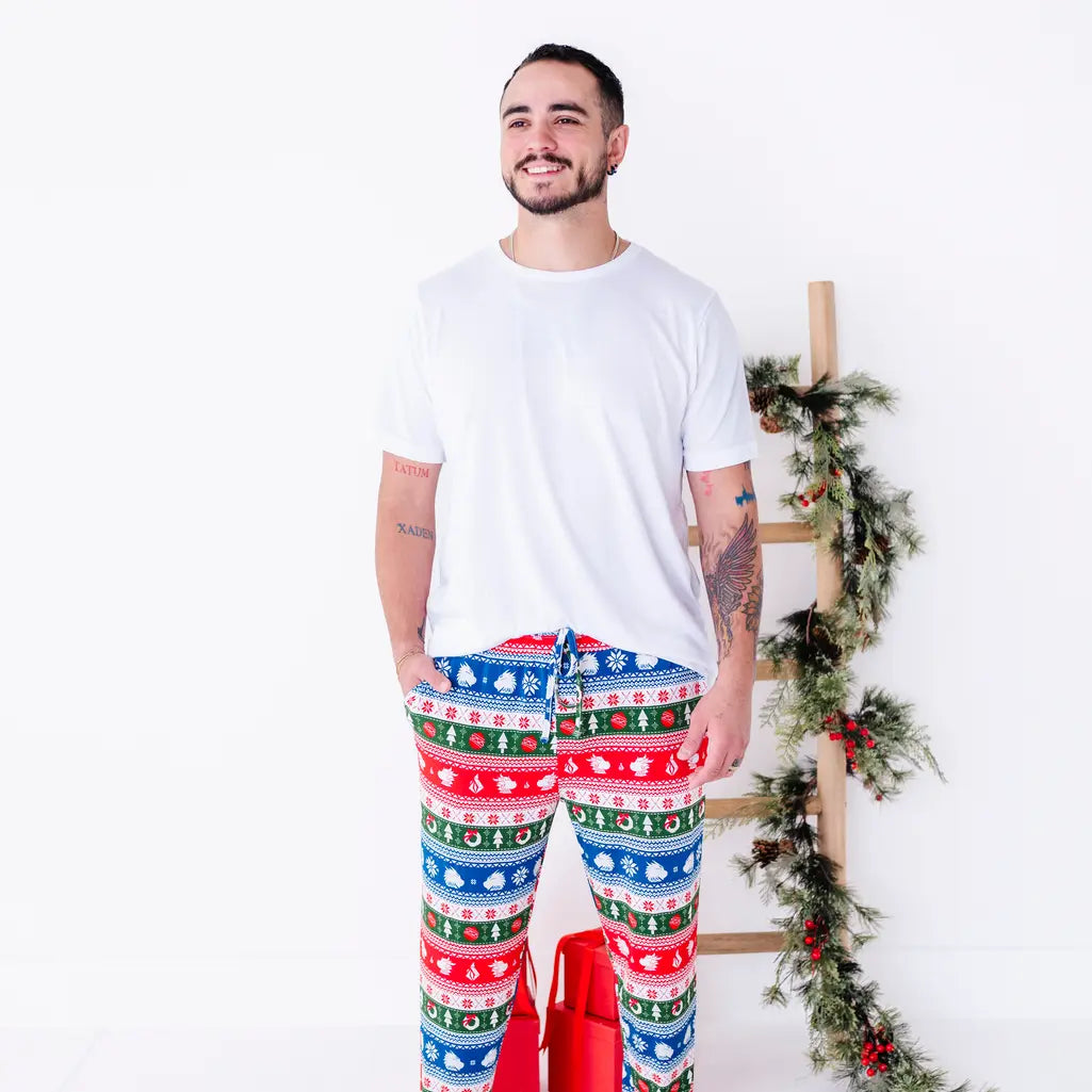 A smiling man in a white tee looking off to the side while standing in jogger pajama pants in red, green, blue, and white fair isle stripes with snowflakes, pine trees, and white dragon heads.