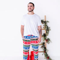 A smiling man in a white tee looking off to the side while standing in jogger pajama pants in red, green, blue, and white fair isle stripes with snowflakes, pine trees, and white dragon heads.