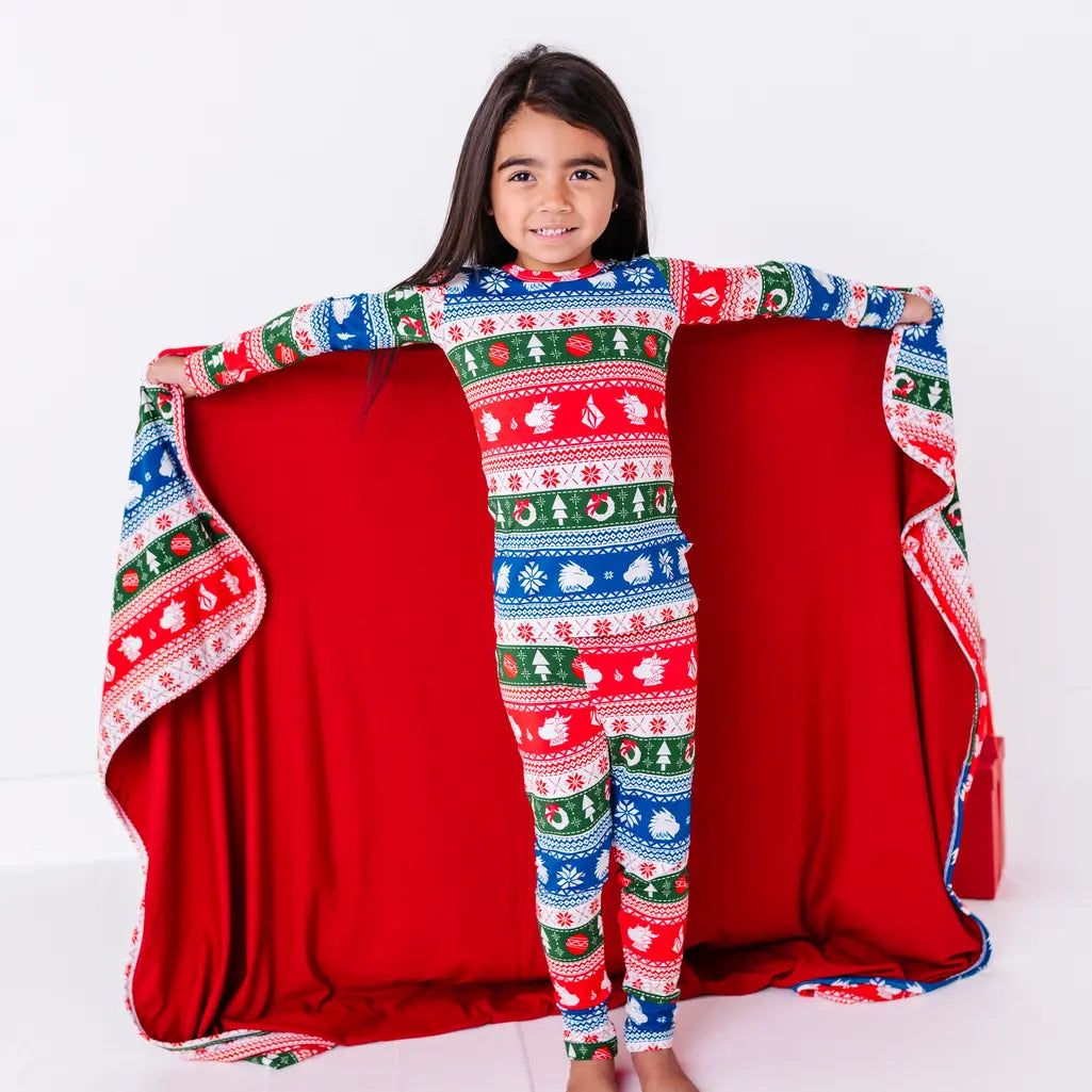 A smiling child holding a blanket in a red, green, blue, and white fair isle print with snowflakes and pine trees open wide to show the red reverse.