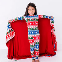 A smiling child holding a blanket in a red, green, blue, and white fair isle print with snowflakes and pine trees open wide to show the red reverse.