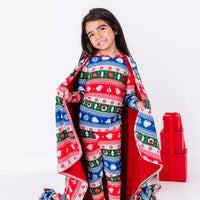 A smiling child with the head tilted, a blanket in a red, green, blue, and white fair isle print with snowflakes and pine trees open like a cape over matching pajamas.