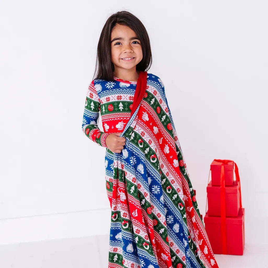 A smiling child standing with a blanket in a red, green, blue, and white fair isle print with snowflakes and pine trees draped over one shoulder and hanging to the floor.