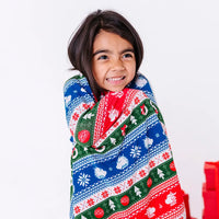 Close up of a child with a big smile wrapped in a blanket in a red, green, blue, and white fair isle print with snowflakes and pine trees.
