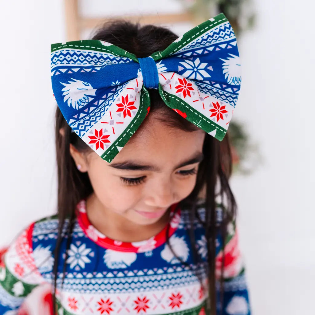 A child looking down while wearing a large fabric bow headband in a red, green, blue, and white fair isle print with snowflakes and white dragon heads, with a matching top.
