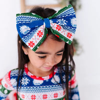 A child looking down while wearing a large fabric bow headband in a red, green, blue, and white fair isle print with snowflakes and white dragon heads, with a matching top.