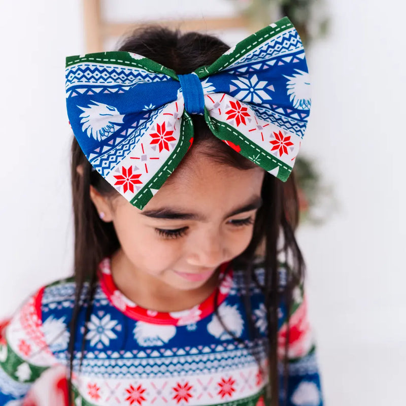 A child looking down while wearing a large fabric bow headband in a red, green, blue, and white fair isle print with snowflakes and white dragon heads, with a matching top.
