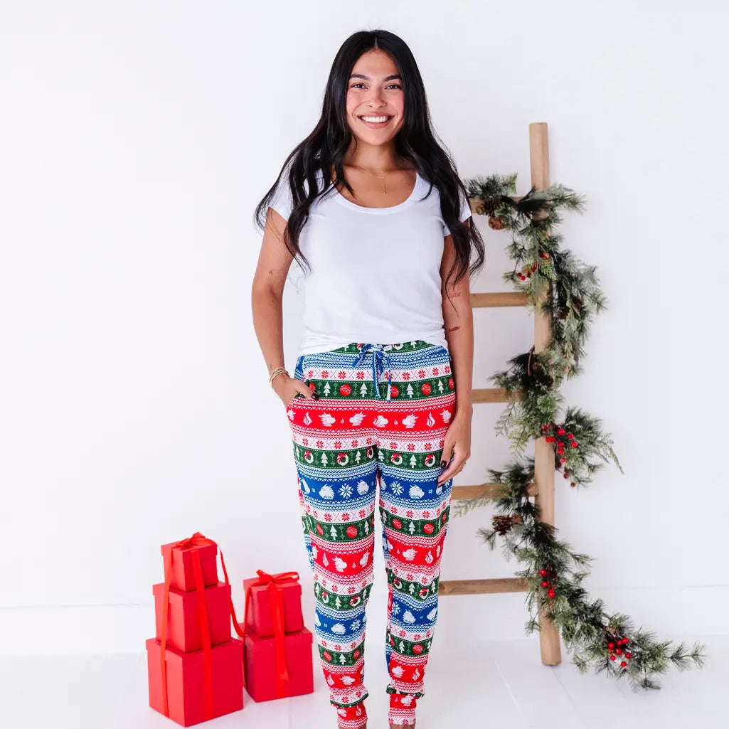 A smiling woman in a white tee standing with a hand in the pocket of jogger pajama pants in red, green, blue, and white fair isle stripes with snowflakes, pine trees, and white dragon heads.