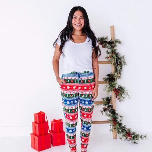 A smiling woman in a white tee standing with a hand in the pocket of jogger pajama pants in red, green, blue, and white fair isle stripes with snowflakes, pine trees, and white dragon heads.