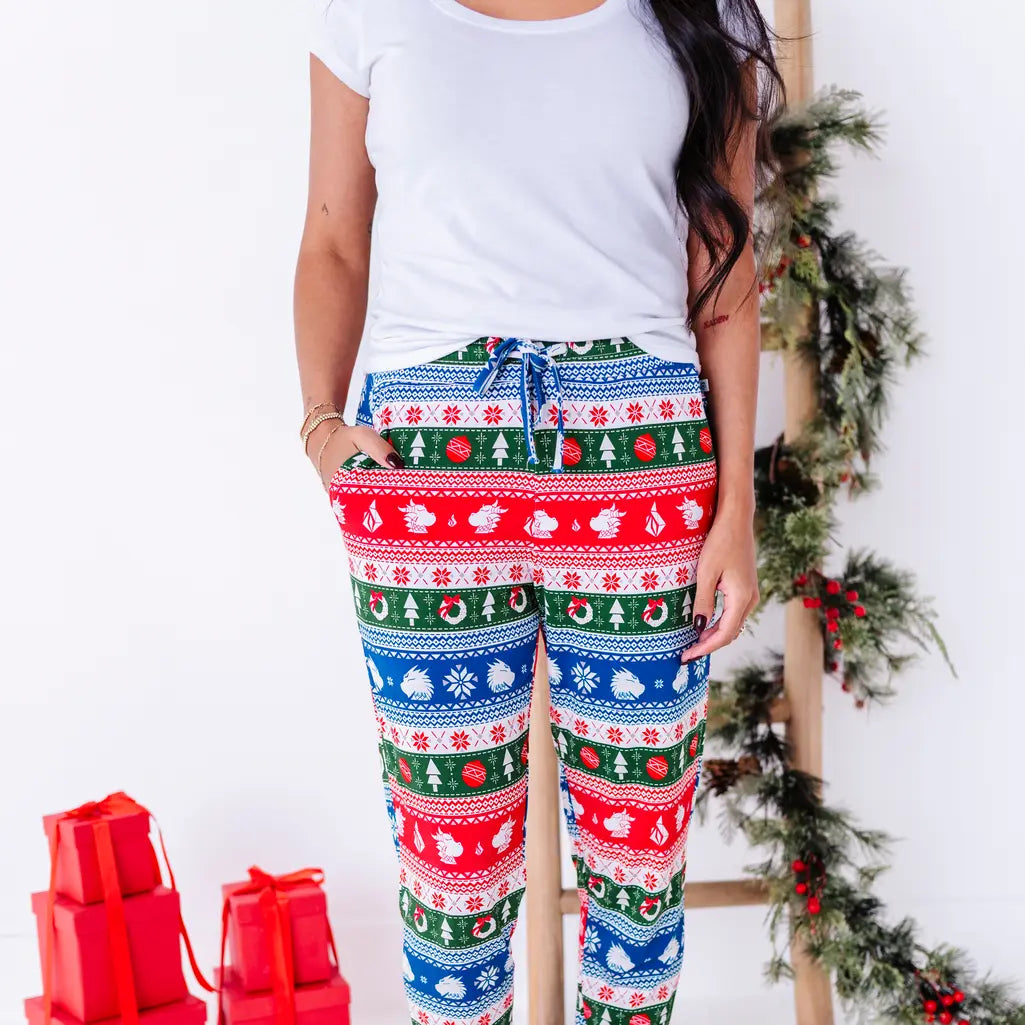 A woman with a hand in the pocket, shown from the chest down, wearing jogger pajama pants in red, green, blue, and white fair isle stripes with snowflakes, pine trees, and white dragon heads.