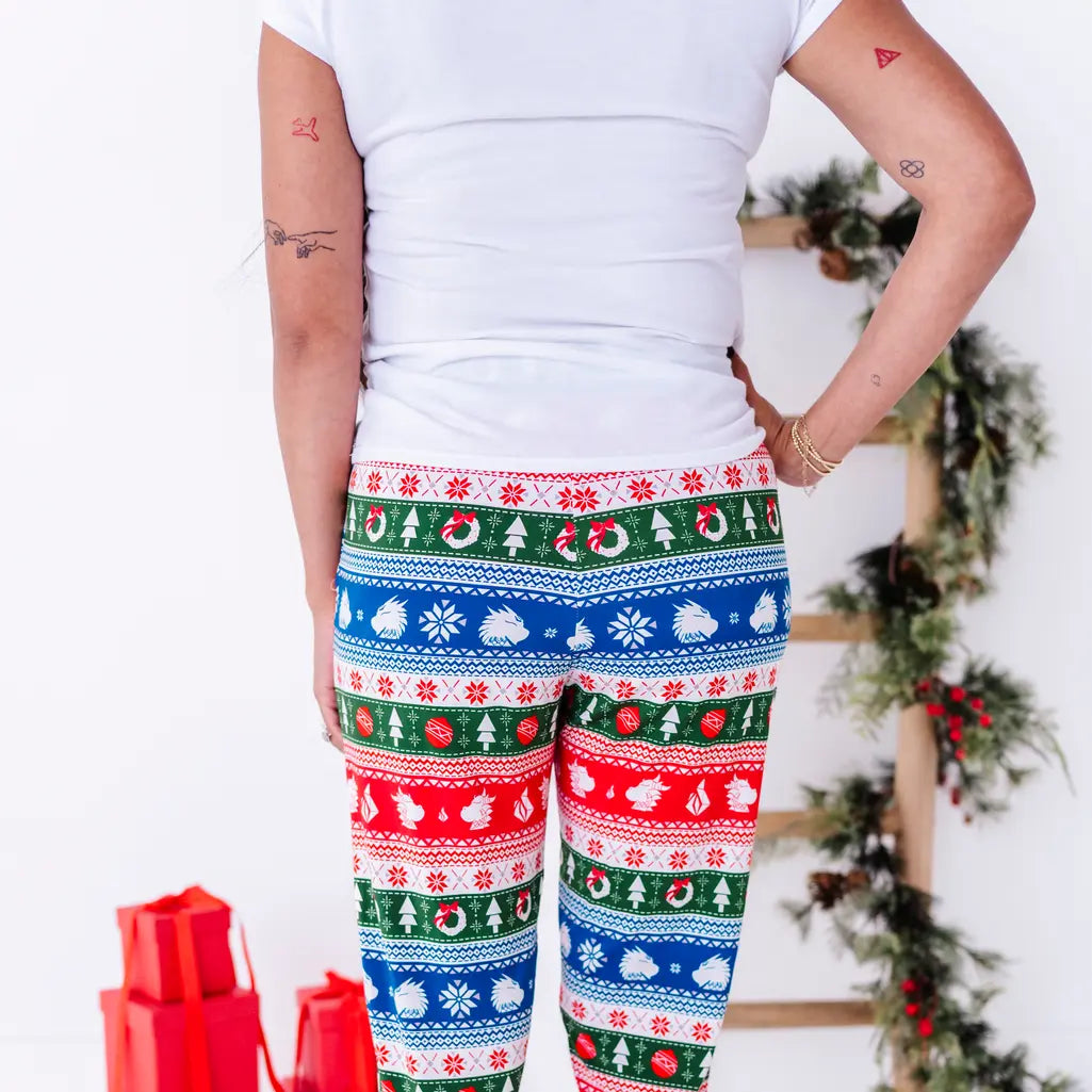 Back view of a woman in a white tee with a hand on the hip, wearing jogger pajama pants in red, green, blue, and white fair isle stripes with snowflakes, pine trees, and white dragon heads.